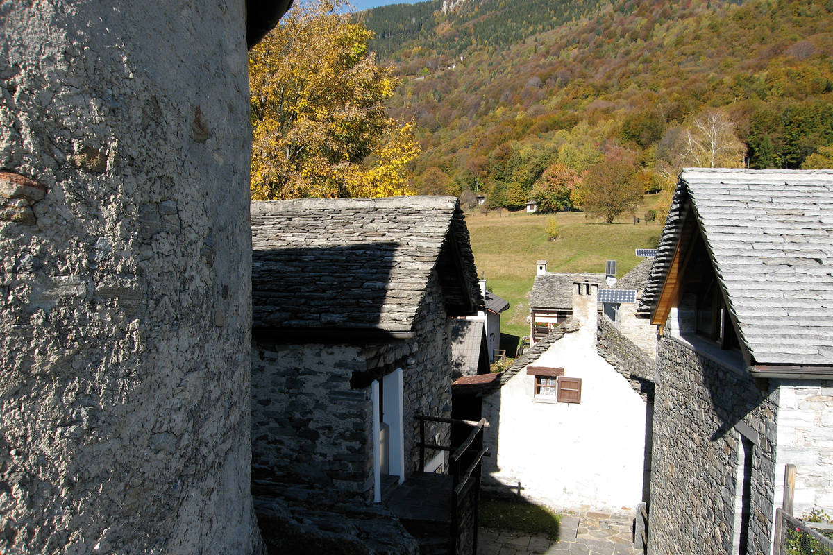 Hinter den typischen, alten Tessiner Steinhäusern erstreckt sich der Wald hügelaufwärts und zeigt sich im Herbst in seinen buntesten Farben.